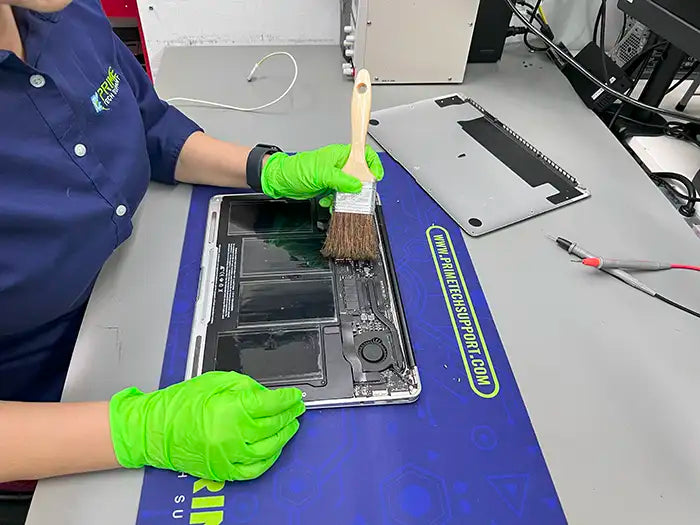 The technician employs a brush to thoroughly cleanse the fans and internal components of the MacBook Air, ensuring optimal performance for a client located in Miami