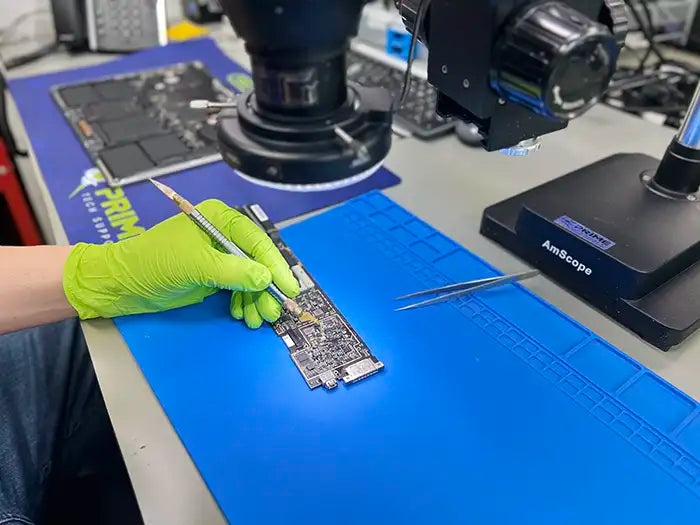 MacBook Air liquid damage repair performed in Prime Tech Support Miami lab. The technician is wearing green gloves and using a scalpel, and looking over a microscope to repair the damage caused by the liquid spill.