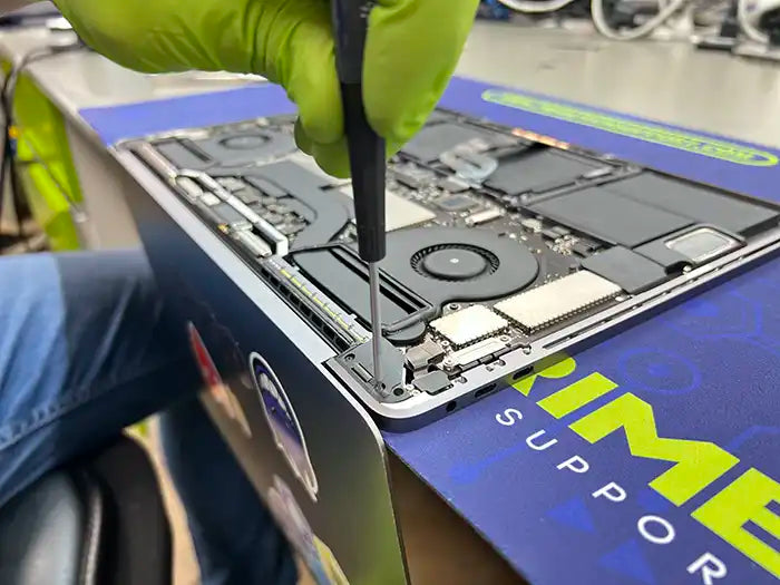 A technician from Prime Tech Support, wearing green gloves, skillfully utilizes a screwdriver to carefully disassemble the rear part of a MacBook Pro. Their purpose is to examine any potential screen damage
