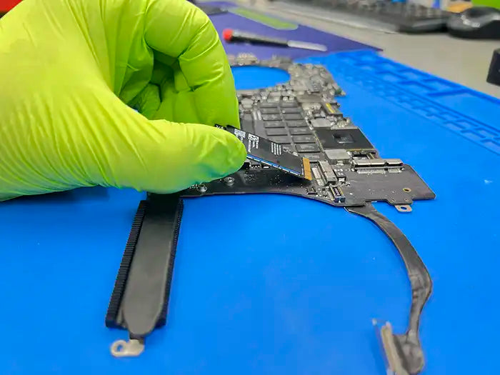 A technician at Prime Tech Support, wearing green gloves, removes a WiFi card from a MacBook Pro to examine and repair it.